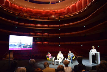 Four people talk and sit on stage in front of an audience with a backdrop of the national opera seats